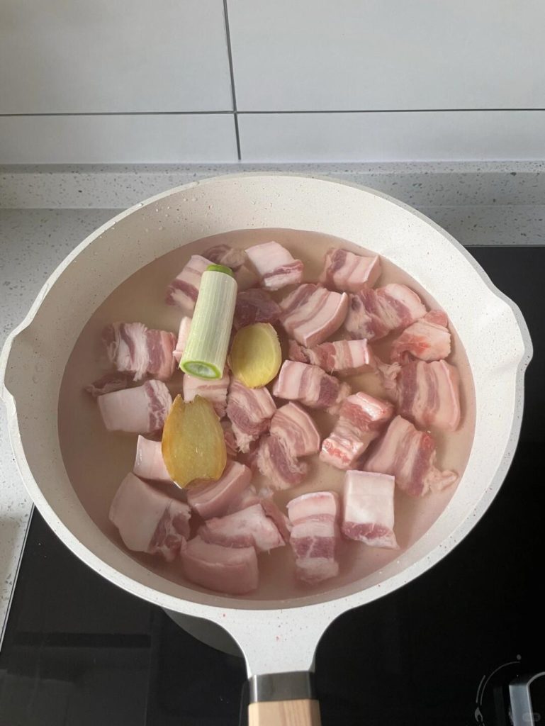 A pot of boiling water with pork belly chunks, ginger slices, and green onion knots inside.
