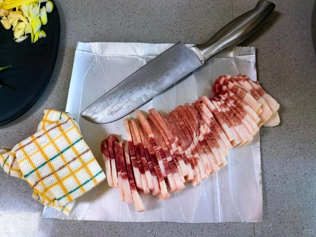 A knife slicing through a block of pork belly that looks slightly frosty/firm.