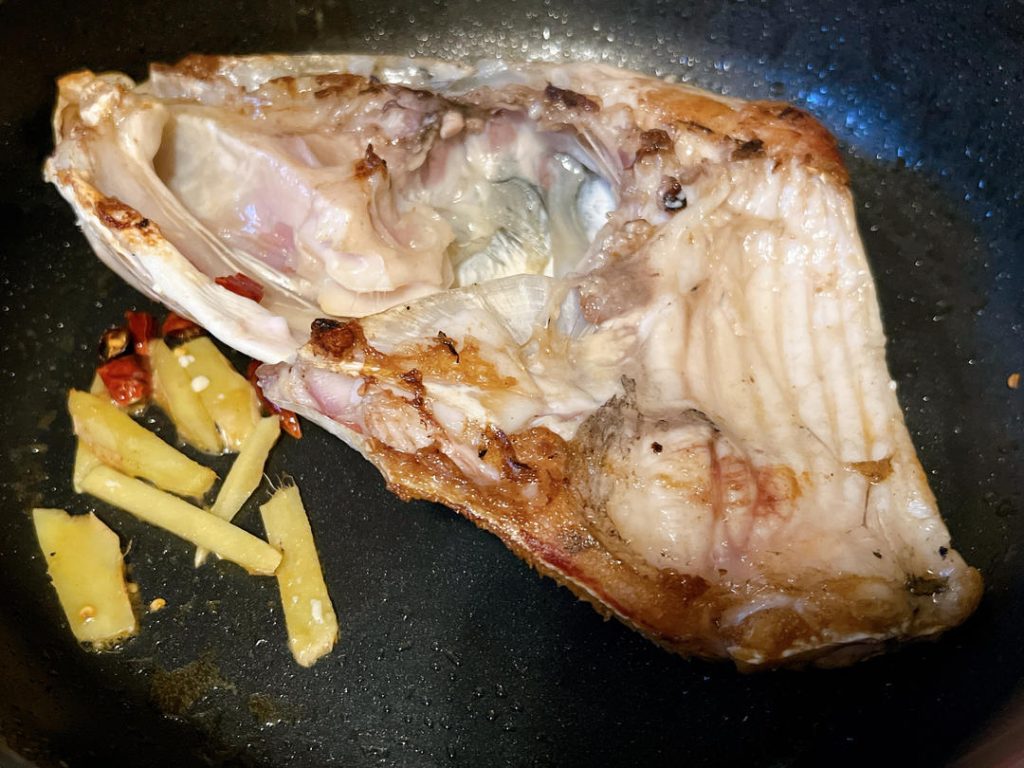 Fry the Aromatics
Gently flip the fish head using a spatula. While the skin side is browning, push the fish slightly to the edge of the wok. Add the ginger slices and optional chilies to the center of the pan. Sauté them in the rendered fish fat for about 30 seconds until highly fragrant.