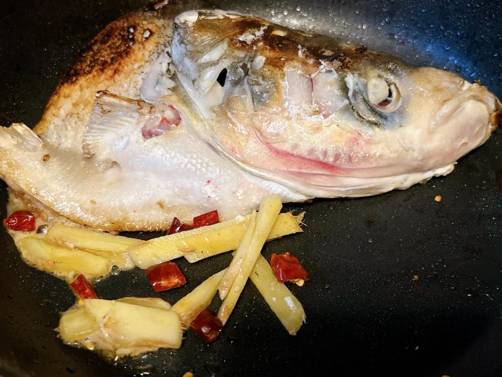 Fry the Aromatics
Gently flip the fish head using a spatula. While the skin side is browning, push the fish slightly to the edge of the wok. Add the ginger slices and optional chilies to the center of the pan. Sauté them in the rendered fish fat for about 30 seconds until highly fragrant.