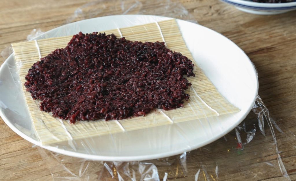 Spread the Rice Scoop a generous amount of the hot purple rice onto the plastic wrap. Wet a spoon (to prevent sticking) and press the rice down into a rectangular shape, about ½ inch (1.2 cm) thick. It needs to be wide enough to encase the fillings.