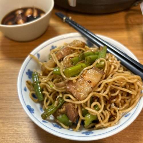 A wide wok filled with Henan braised noodles, pork belly, and green beans, steam rising from the glossy noodles