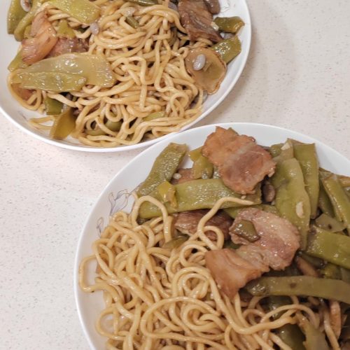 Braised noodles with green beans and pork belly in a wok, tossed together and steaming