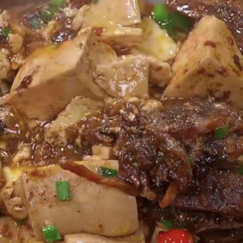 Braised tofu with dace in black bean sauce in a white dish, topped with sliced red chilies and scallions