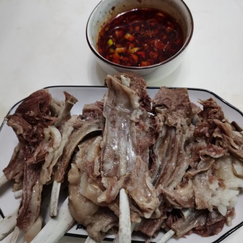 Whole boiled lamb rack on a cutting board with a bowl of chili dipping sauce