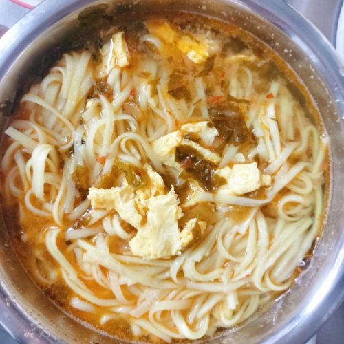 A bowl of sour soup egg noodles with fluffy scrambled eggs, pickled greens, and chili oil