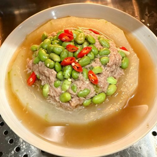 Steamed winter melon with pork and salted egg yolk topped with edamame and scallions on a white plate